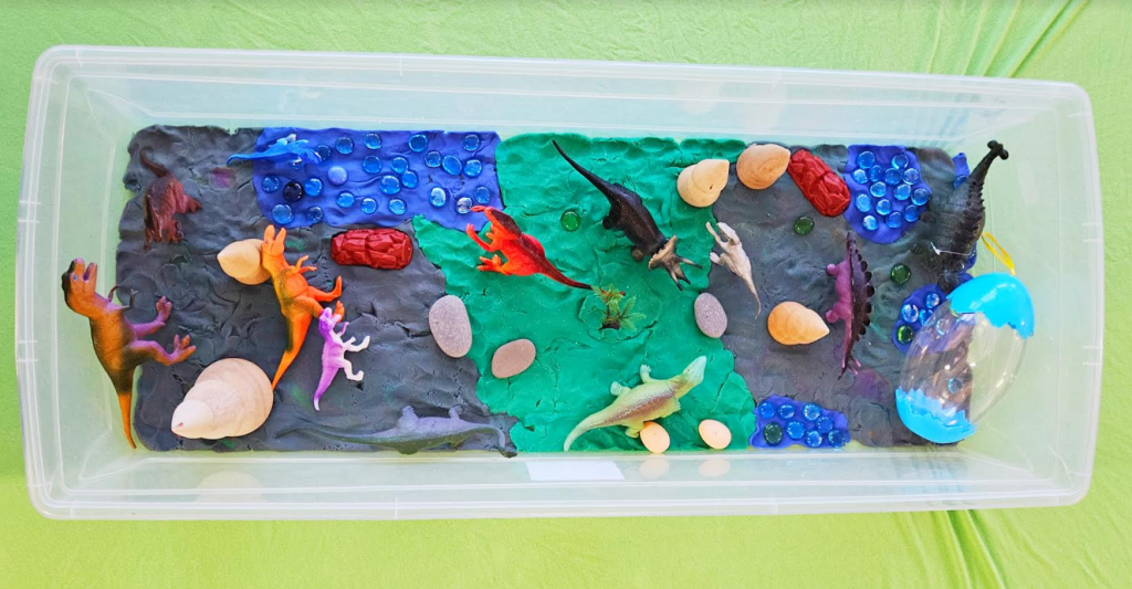 sensory play for kids shows a bin with green and blue play dough and dinos, rocks and beads.