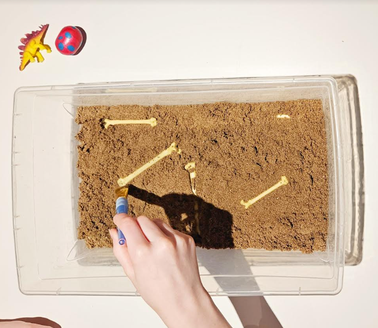 dinosaur activities for kids shows a child using a paint brush to brush off bones in a bin with sand.
