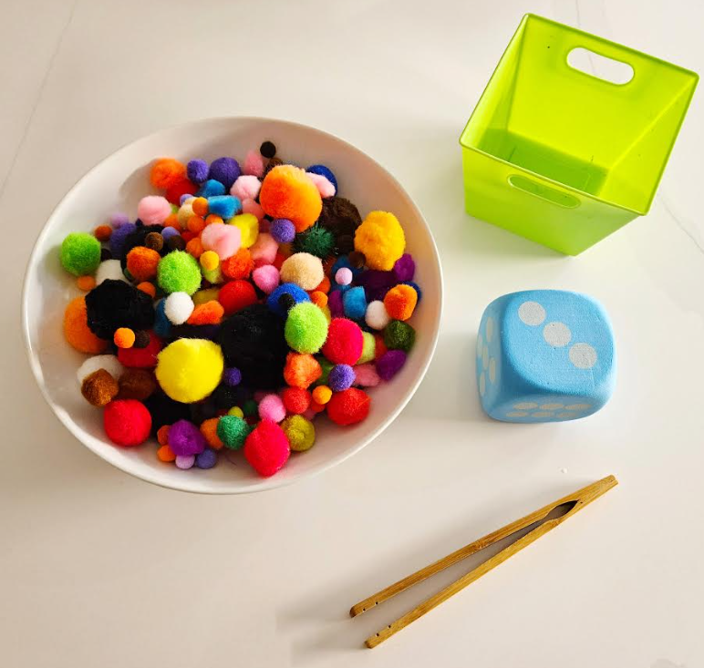Easy Kindergarten Math Activity shows a bowl of pompoms, a die, square container and chopsticks.