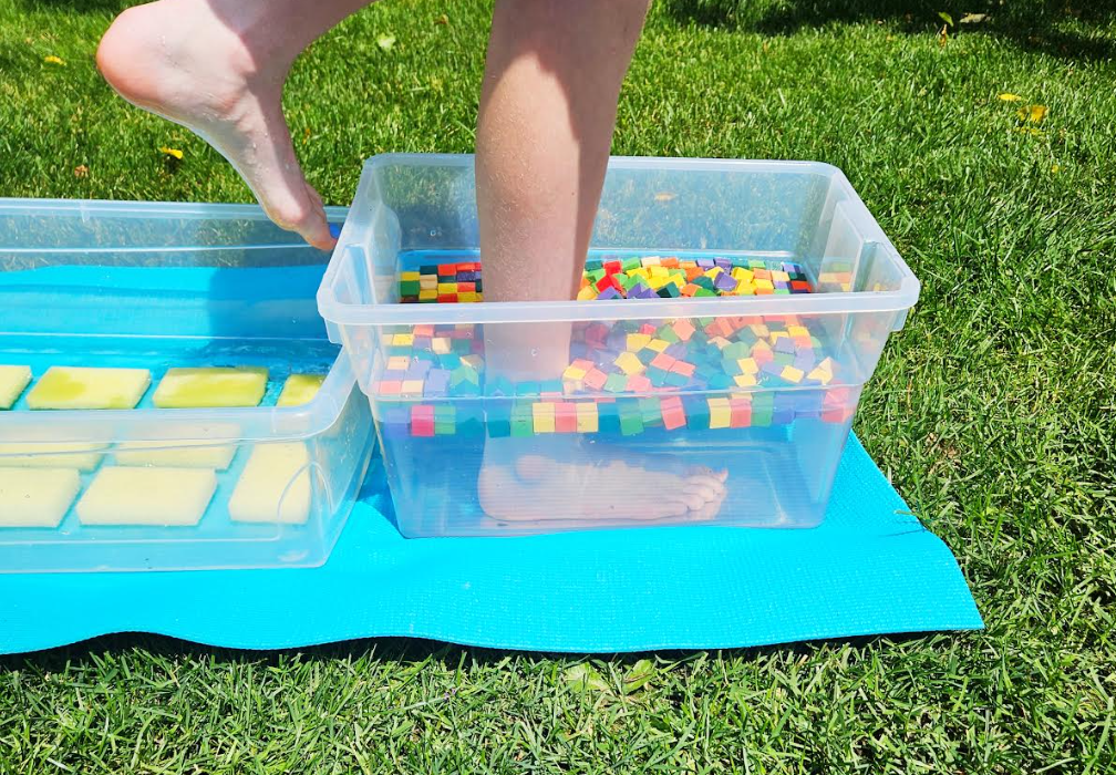 outdoor summer activity shows a child stepping into a bin filled with water and wooden cubes.