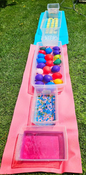 summer sensory path shows a row of bins with different summery items like water balloons, ice etc.