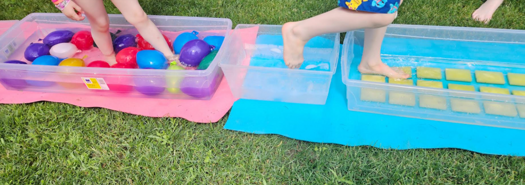summer sensory path shows children walking through bins filled with sponges and water balloons.