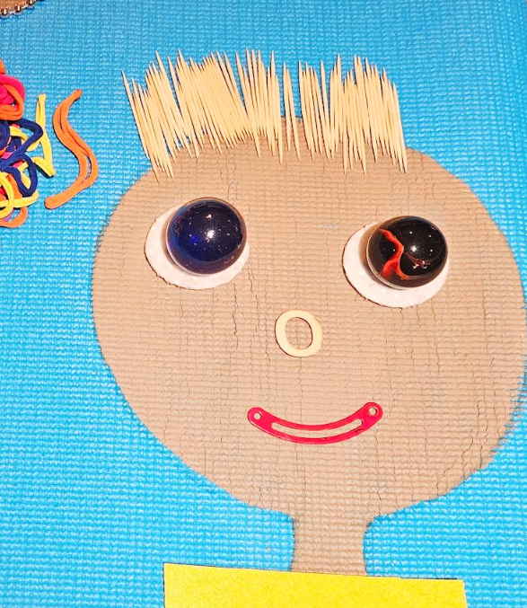 crafts for kids shows a face made on a yoga mat that has hair with toothpicks and featured from loose parts.